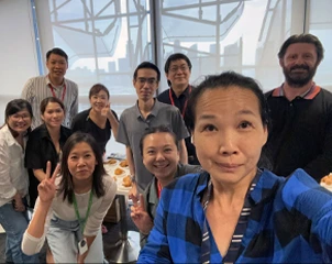 Selfie grupal de compañeros de trabajo reunidos alrededor de una mesa con comida, sonriendo y haciendo el signo de la paz.